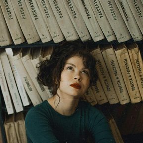 Une jeune femme brune, les cheveux noués négligemment, les lévres peintes d'un rouge pourpre, vêtue d'un pull vert sapin, est assise au pied d'une bibliothèque dont tous les livres sont beiges avec une écriture noire sur la tranche, illustrant le travail.