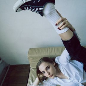 Une jeune femme blonde affalée sur un canapé en mousse beige. Elle porte un chemisier blanc, un pantalon noir, des chaussettes blanches à mi-cheville et des tennis noires et blanches. Elle lève nonchalement une jambe au-dessus d'elle, telle une gymnaste. 