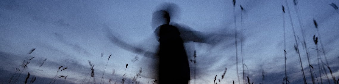 Photo poétique prise en slow motin d'une personne en mouvement, dansant peut-être, entre de hautes herbes, au moment des vespres, alors que les dernières lueurs du jour disparaissent à l'horizon. Le ton de la photo est bleuté et inspire la douceur. 