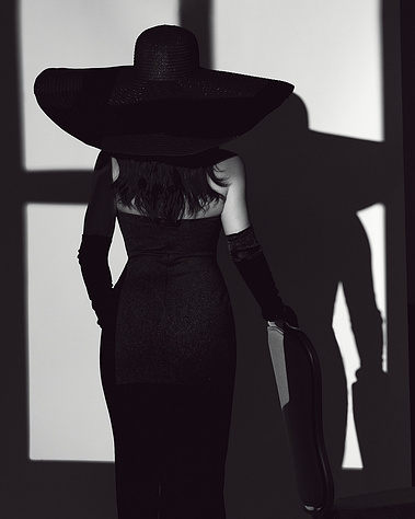 Black and white portrait of a woman in a fitted gown, wide-brim hat, and black gloves, standing in front of a dramatic light source. Shadows and contrast enhance this artistic editorial-style image, ideal for fine art and fashion photography in Arlington 