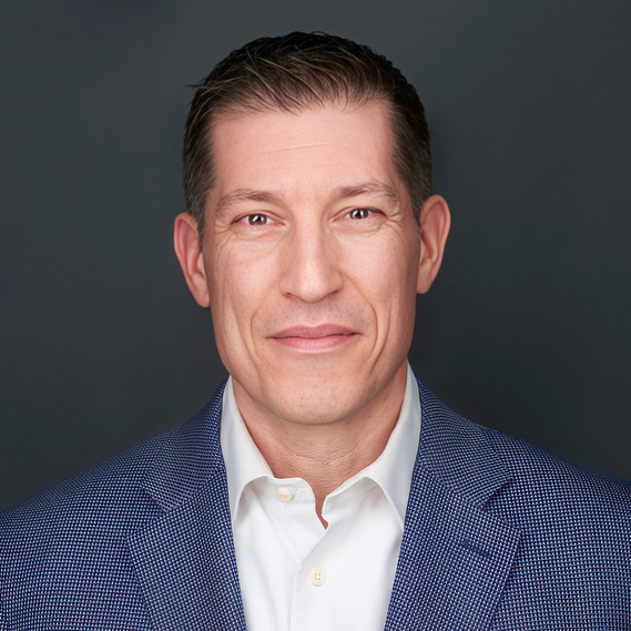 Corporate headshot of a man with short dark hair, wearing a white dress shirt and a blue textured blazer against a neutral gray background. Ideal for executive branding, LinkedIn profiles, leadership portraits, and business networking in Arlington Heights