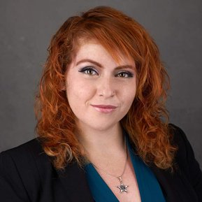 Professional headshot of a woman with red curly hair wearing a navy blazer and blue top against a dark gray background. Ideal for business branding, corporate profiles, and LinkedIn in Arlington Heights and Chicago.