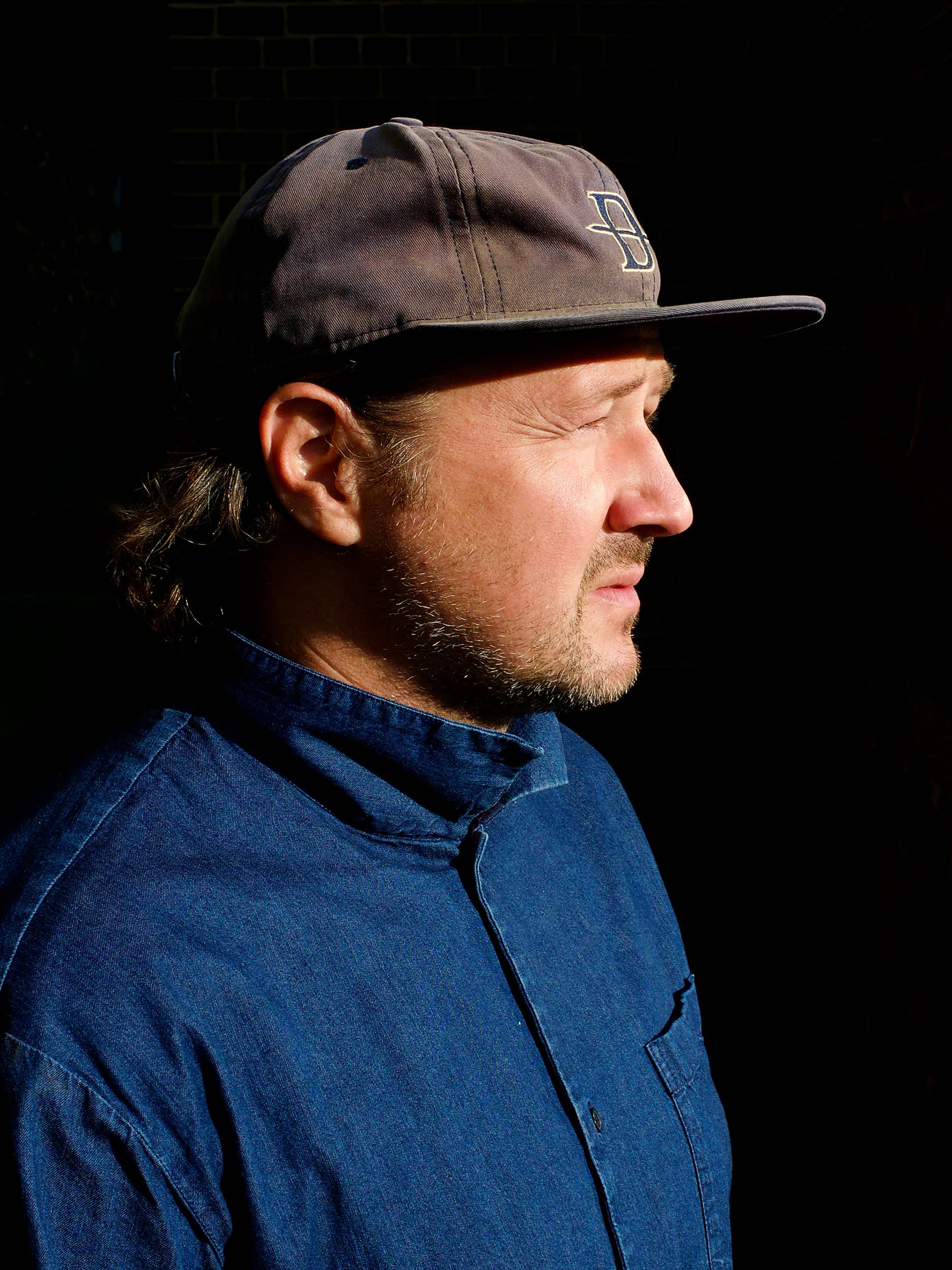 Man stands in the sun wearing a denim shirt and a grey Daneson cap. 