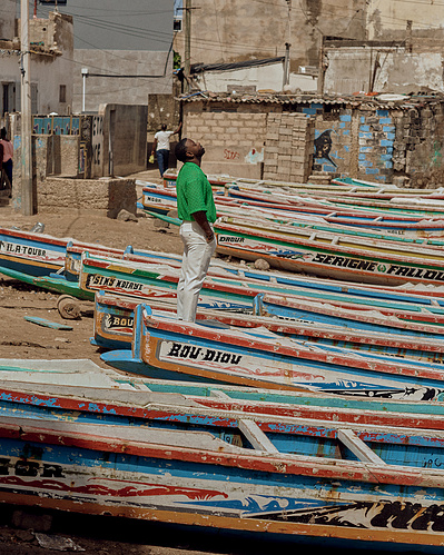 Steven DK Brown with Bryan Michael Ham Photography, at Petit Ngor Beach in Dakar, Senegal. Basking in the African Heat by the colorful fishing boats. wearing a green top, and white pants. 