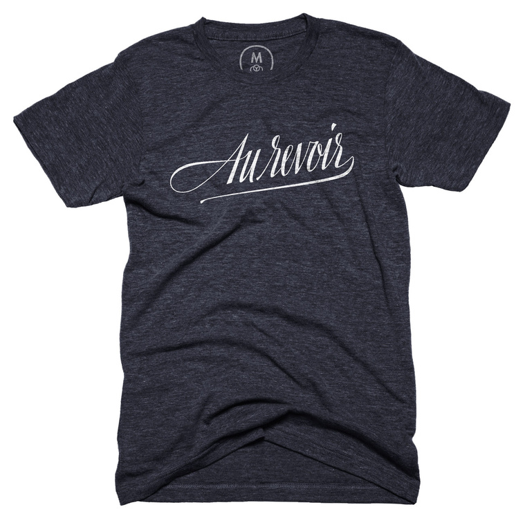 A navy blue tshirt with the French phrase “Au Revoir” (“goodbye” in English) neatly custom lettered in a compressed italic script, in the style of a calligraphy nib pen, in white ink.