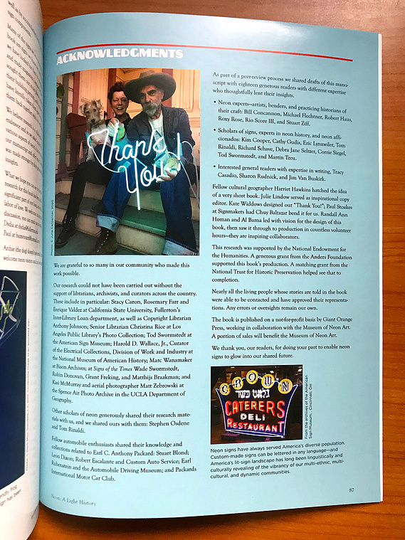 Page in a book featuring a photo of a pair of authors and their small dog sit on the porch of their house, holding a lit aquamarine blue neon sign that says 