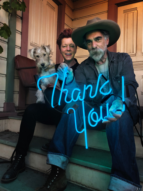 A pair of authors and their small dog sit on the porch of their house, holding a lit aquamarine blue neon sign that says 