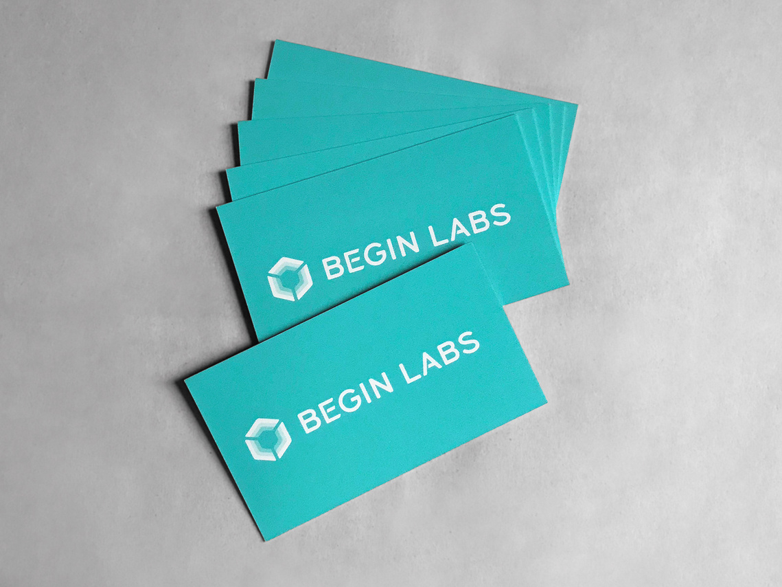 The Begin Labs logo design, using a custom lettered wordmark, featured on a printed stack of bright turquoise business cards. 