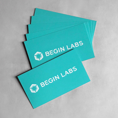 The Begin Labs logo design, using a custom lettered wordmark, featured on a printed stack of bright turquoise business cards. 