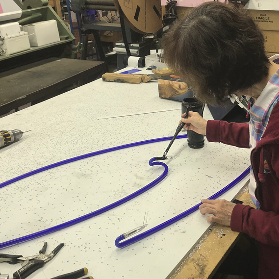 A woman at Artico Lite applies blackout paint to the ends of a blue neon tube bent in the design of script lettering. 