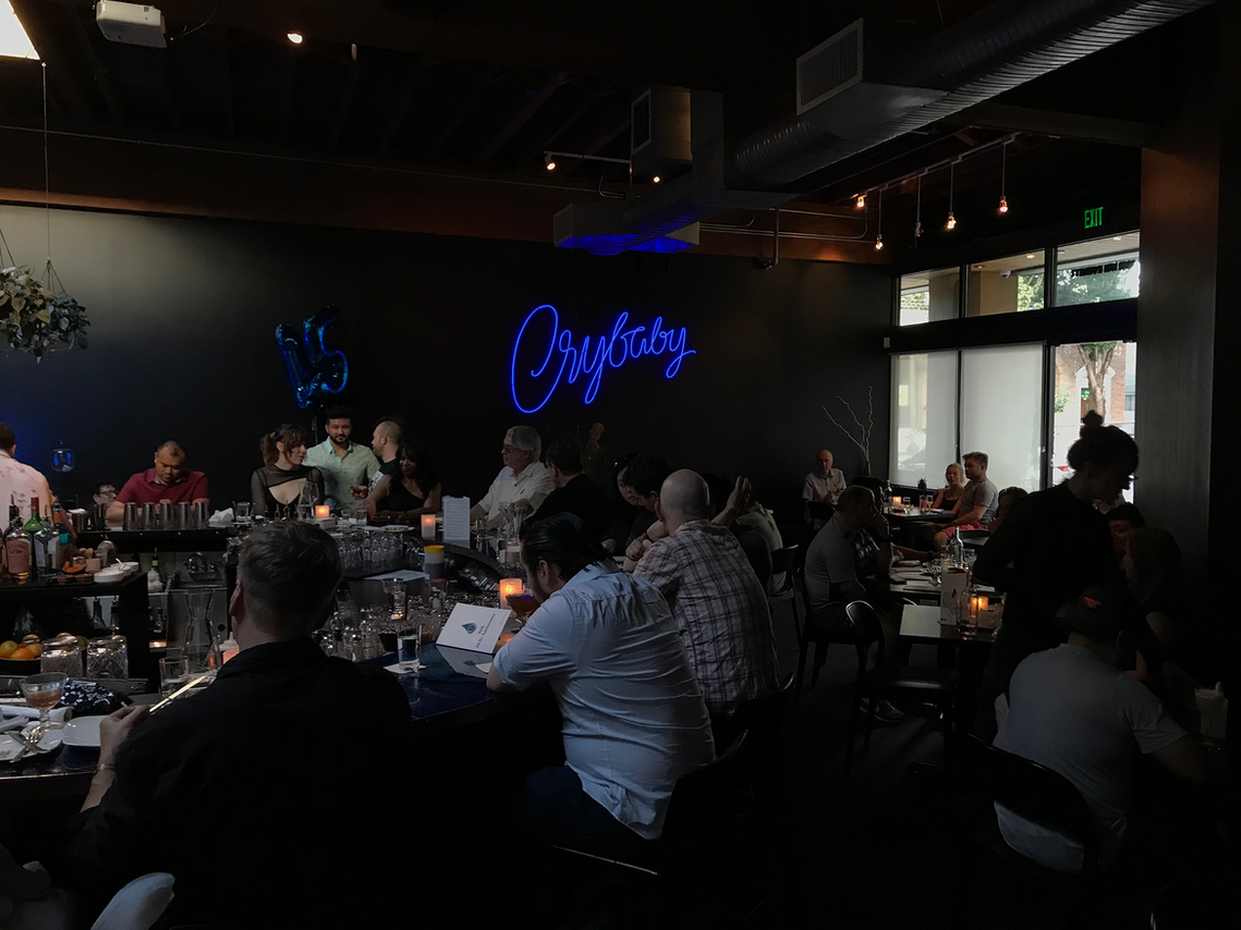 A busy bar scene with people sitting and standing, while a lit neon sign, designed with expressive blue script lettering, spelling out the word 