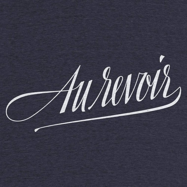 Closeup of a navy blue tshirt with the French phrase “Au Revoir” (“goodbye” in English) neatly custom lettered in a compressed italic script, in the style of a calligraphy nib pen, in white ink.