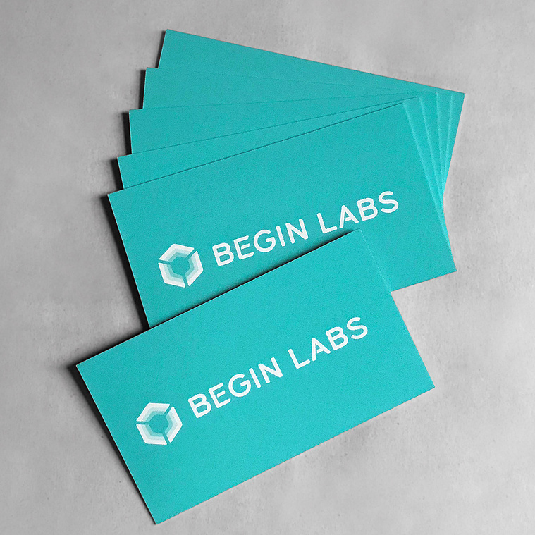 The Begin Labs logo design, using a custom lettered wordmark, featured on a printed stack of bright turquoise business cards. 