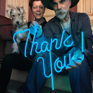 A pair of authors and their small dog sit on the porch of their house, holding a lit aquamarine blue neon sign that says 