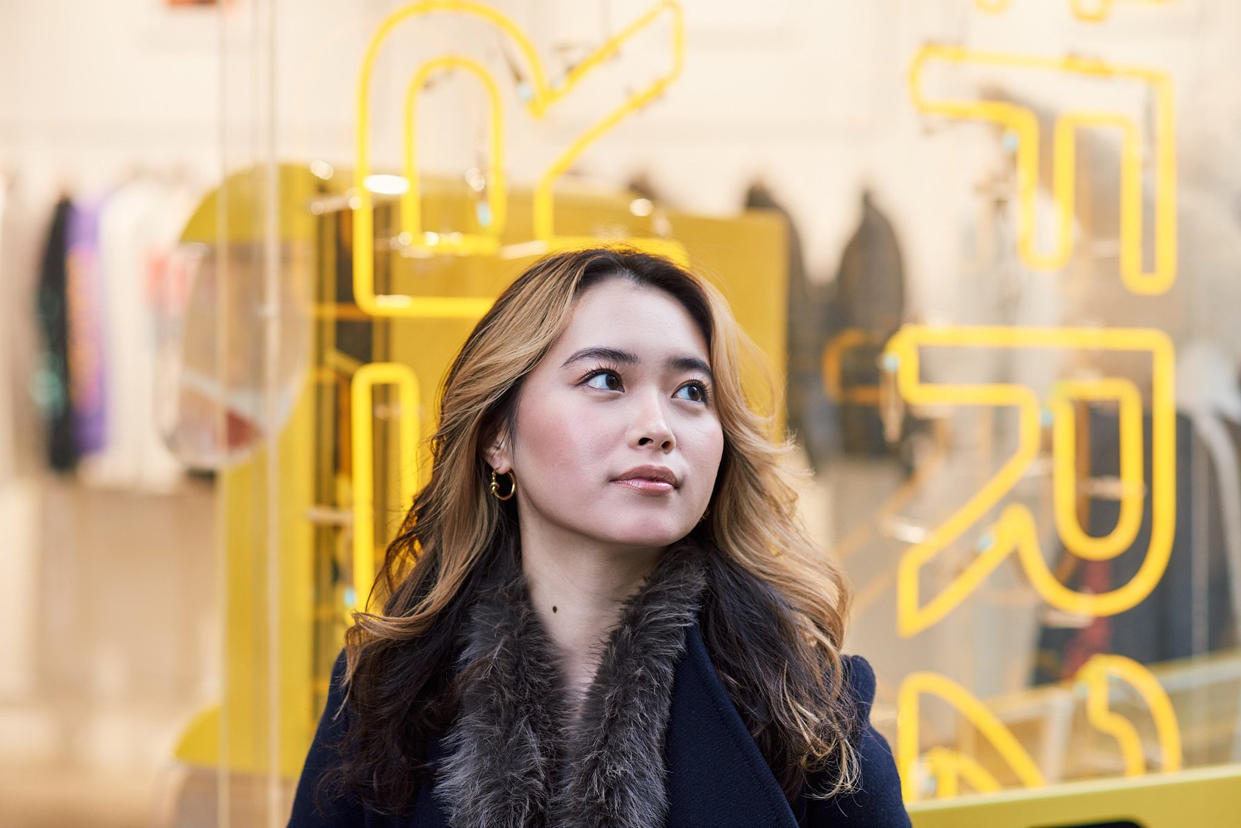 Portrait of a Japanese woman, Minori Abe, with blonde highlights in her hair, standing in front of a store window with a yellow neon sign.