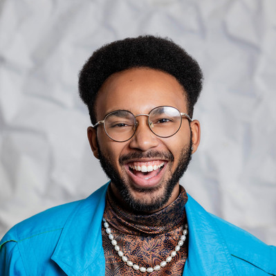 A Corporate headshot of a smiling black man with a short afro, a beard, wearing black glasses, a black paisley turtleneck ,and a bright blue blazer.
http://www.annanproductions.com/