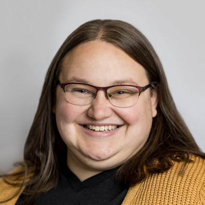 A Corporate headshot of a smiling white woman with straight medium-length brown hair, wearing brown glasses, a yellow knitted sweater, and a black inner t-shirt. 
http://www.annanproductions.com/