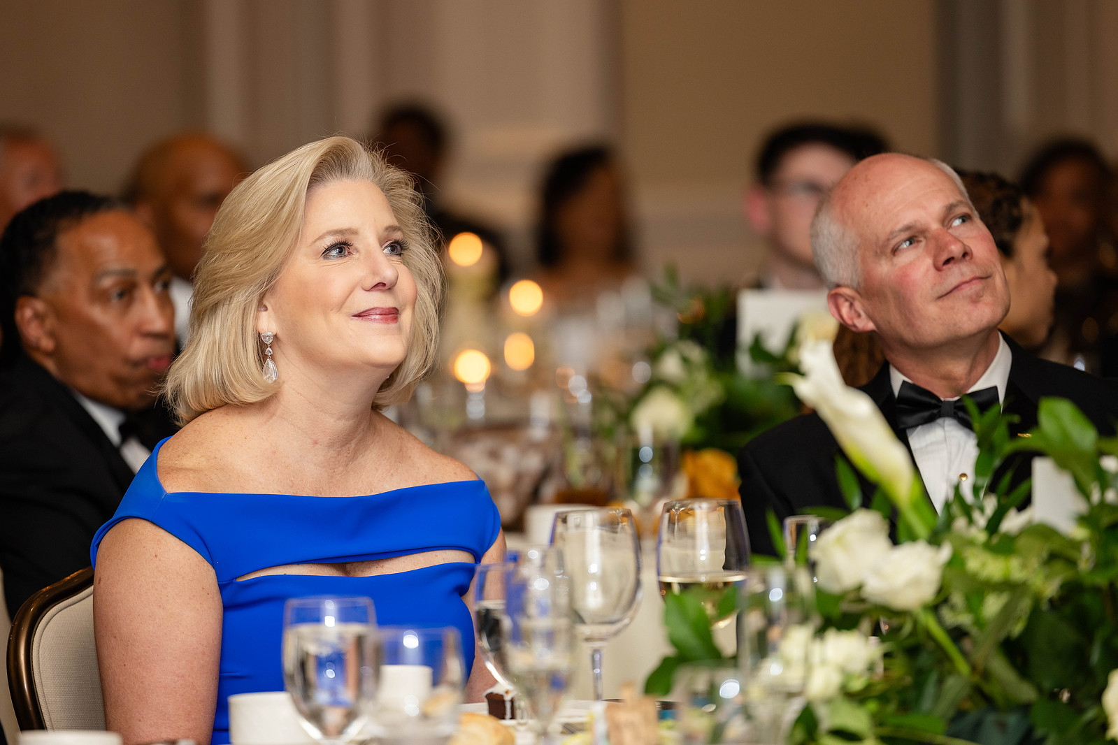 ROCKS INC 2024 Spring Gala. Hon. Christine Wormuth, Secretary sitting at a dinner table with white roses, observing and listening to someone speaking. Annan Productions LLC. www.annanproductions.com