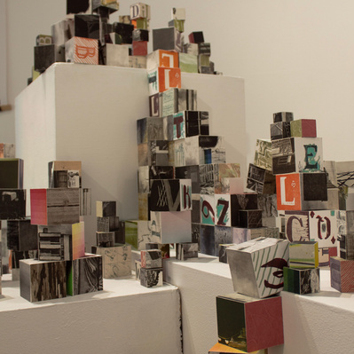 An installation of hundreds of blocks of various sizes stacked on top of white plinths. Each block has printed imagery adhered to each side.