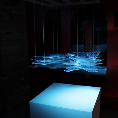 A floating block of transparent sheets in various shapes, with engraved lines of maps and architectural plans on them. They are illuminated by a dramatic blue light and hang over a flat white surface.