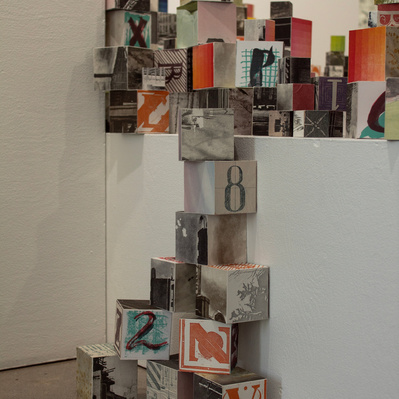An installation of hundreds of blocks of various sizes stacked on top of white plinths. Each block has printed imagery adhered to each side.