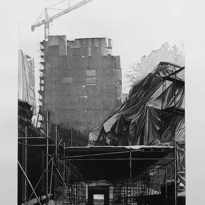 A black and white illustration of a scaffolding tunnel, leading to a bright point of light. Above the tunnel, two large, looming tarp-covered structures dominate the composition.