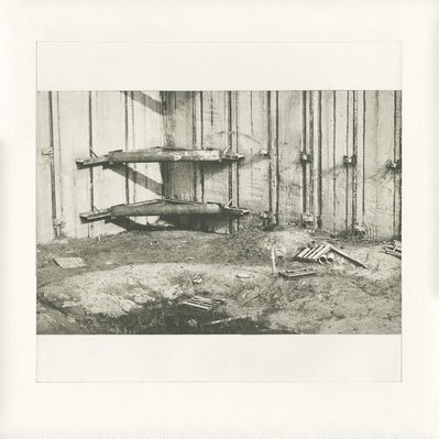 A deep construction excavation, with several corner braces dominating the centre, and shipping palettes and broken wood in the mud at the bottom.