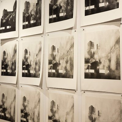 A group of prints on a white gallery wall. They all depict the same structure wrapped in tarps,  but in each print read left to right, top to bottom, the image becomes more and more worn away.