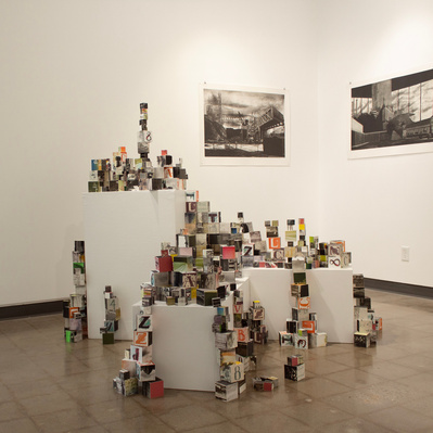 An installation of hundreds of blocks of various sizes stacked on top of three white plinths, in front of two black and white etchings. The blocks have images printed on all of their sides.
