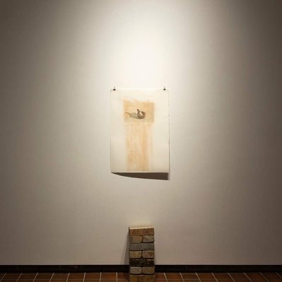 A neat stack of bricks, two wide, stands on the gallery floor. Behind it, a photographic print is tacked to the wall, covered with brick-red dust.