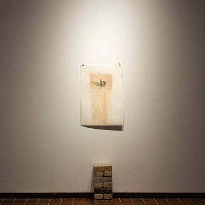 A neat stack of bricks, two wide, stands on the gallery floor. Behind it, a photographic print is tacked to the wall, covered with brick-red dust.