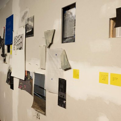 An unfinished wall, with the drywall seam visible. A selection of monochromatic prints of parking garages and construction sites are arrayed across it somewhat randomly, with yellow post-it notes, fragments of tarp, Tyvek, and thin cast concrete.