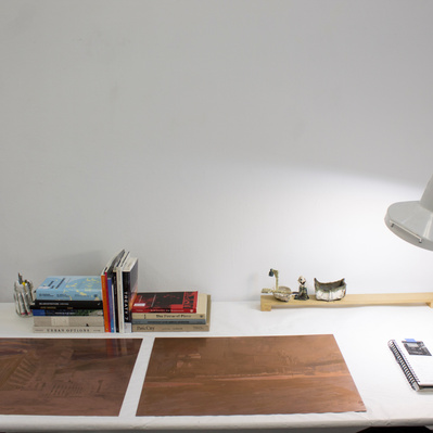 A desk with two large sheets of copper, a sketchbook, several books, a small ceramic boat, and a lamp.