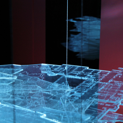 A floating block of transparent sheets in various shapes, with engraved lines of maps and architectural plans on them. They are illuminated by a dramatic blue light and are reflected in a window behind them in the dark space.