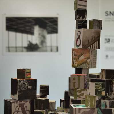 An installation of hundreds of blocks of various sizes stacked on top of white plinths. Each block has printed imagery adhered to each side.