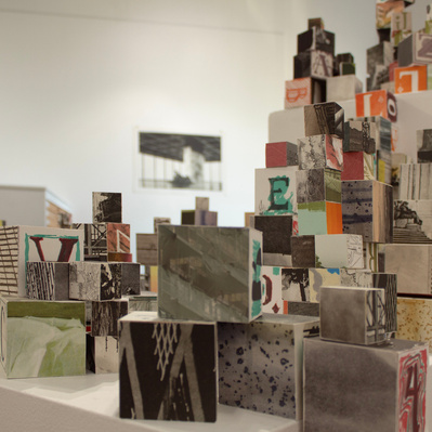An installation of hundreds of blocks of various sizes stacked on top of white plinths. Each block has printed imagery adhered to each side.