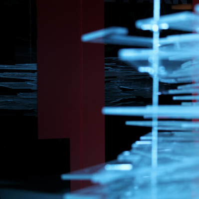 A floating block of transparent sheets in various shapes, with engraved lines of maps and architectural plans on them. They are illuminated by a dramatic blue light and are reflected in a window behind them in the dark space.
