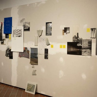 An unfinished wall, with the drywall seam visible. A selection of monochromatic prints of parking garages and construction sites are arrayed across it somewhat randomly, with yellow post-it notes, fragments of tarp, Tyvek, and thin cast concrete.