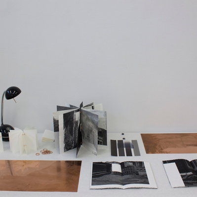 A table with two large sheets of copper, a lamp, and several open book spreads in with black and white images visible.
