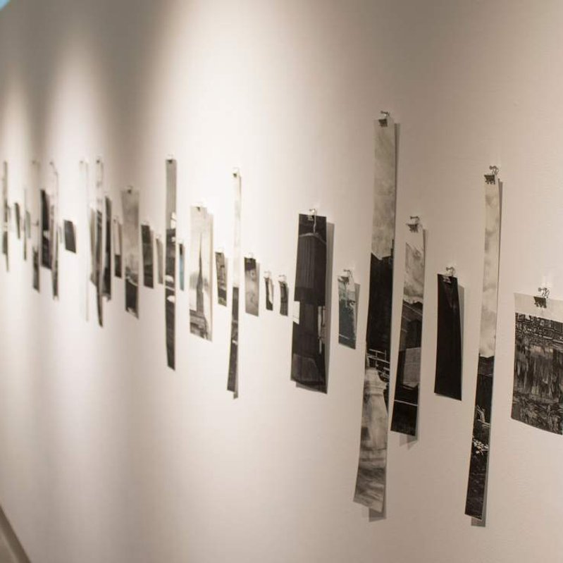 On the white wall of a gallery, a series of tall, narrow strips of prints of varying heights are clipped to the wall, evenly spaced.