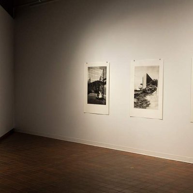 A room in a gallery, with white walls and red-orange tiled floors. Two walls facing each other are visible, and there are three black and white prints on each wall.