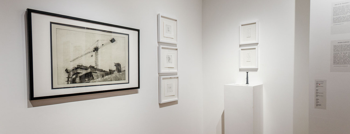 A gallery installation, featuring a large print of a crane, three small prints in white frames hung vertically, a plinth with a tiny black column on it, and two framed pieces above it.