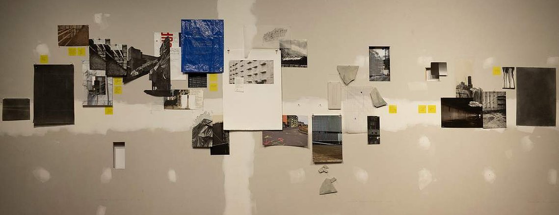 An unfinished wall, with the drywall seam visible. A selection of monochromatic prints of parking garages and construction sites are arrayed across it somewhat randomly, with yellow post-it notes, fragments of tarp, Tyvek, and thin cast concrete.
