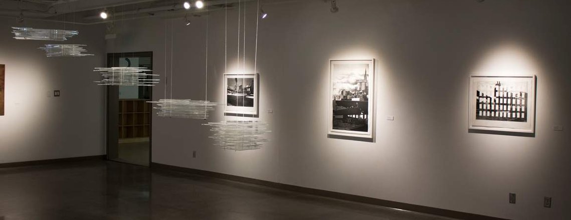 Several architectural sculptural components made from layers of horizontal transparent layers of acrylic are hanging in the centre of a gallery space, with three framed artworks on the wall behind.