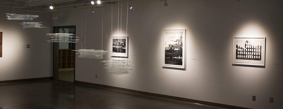 Several architectural sculptural components made from layers of horizontal transparent layers of acrylic are hanging in the centre of a gallery space, with three framed artworks on the wall behind.