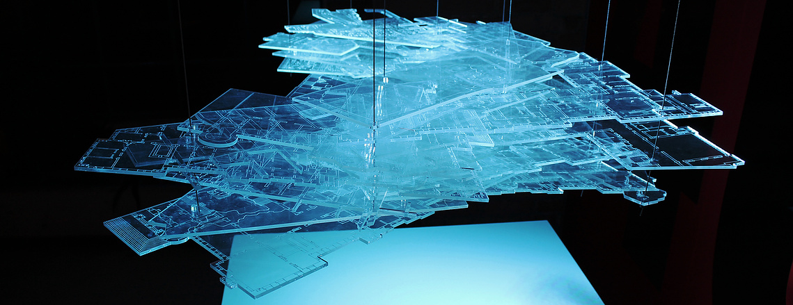 A floating block of transparent sheets in various shapes, with engraved lines of maps and architectural plans on them. They are illuminated by a dramatic blue light and hang over a flat white surface.