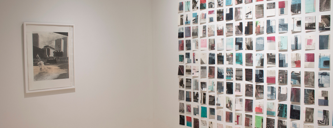 A grid of small prints on the right, and a single framed larger print on the left, are hung on the white walls of a gallery.