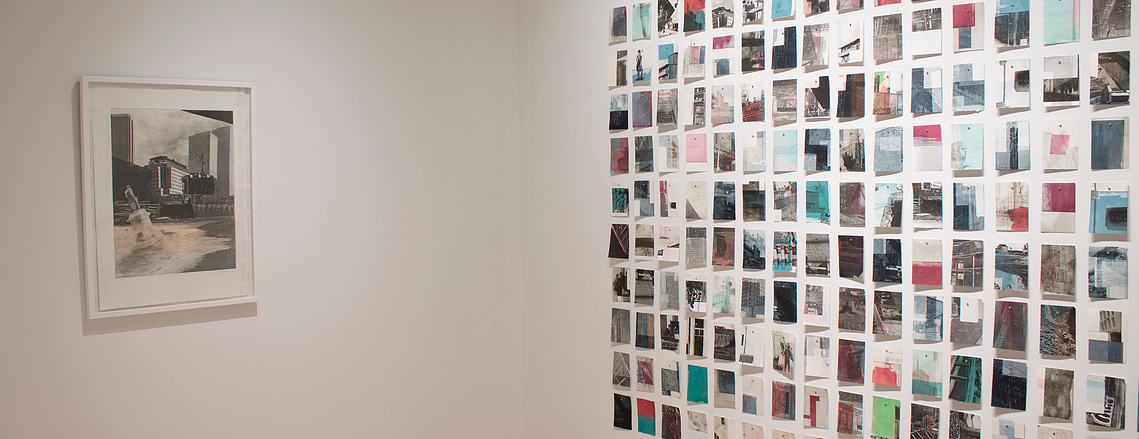 A grid of small prints on the right, and a single framed larger print on the left, are hung on the white walls of a gallery.