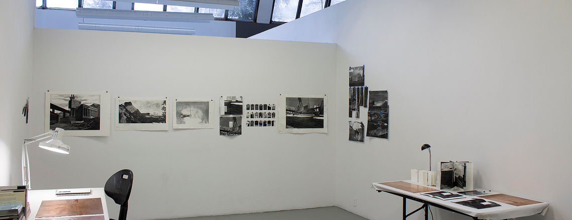 A studio with white walls and clerestory windows is furnished with two tables, facing each other on opposite walls. The wall between them has a series of black and white prints pinned up, while sheets of copper and other objects sit on the tables.