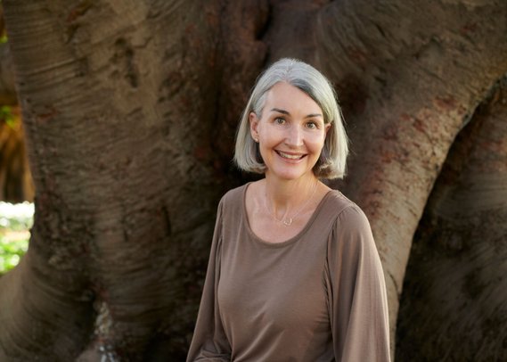 photo of a lady in a brown shirt with grey hair. There is a tree trunk in the background